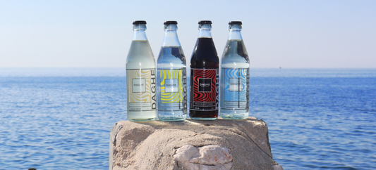 Quatre bouteilles de boissons Daghe : Tonic, Ginger Beer, Cola et Limonade, posées sur un rocher face à la mer, avec l'horizon et les vagues en arrière-plan.