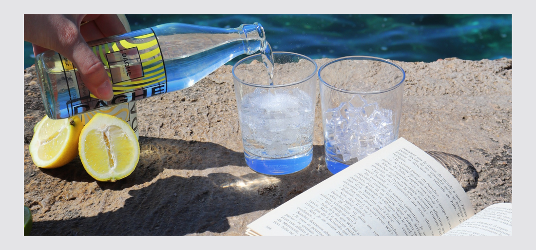 Vue mer avec verre rempli de glaçons, bouteille DAGHE Tonic versée dans un des deux verres, livre ouvert posé devant.