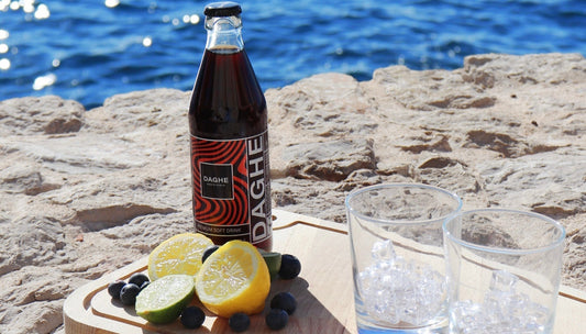 Bouteille de Daghe Monte Carlo version cola avec étiquette rouge posée sur une planche en bois, accompagnée de deux verres avec des glaçons. À côté des citrons et des myrtilles, en bord de mer.